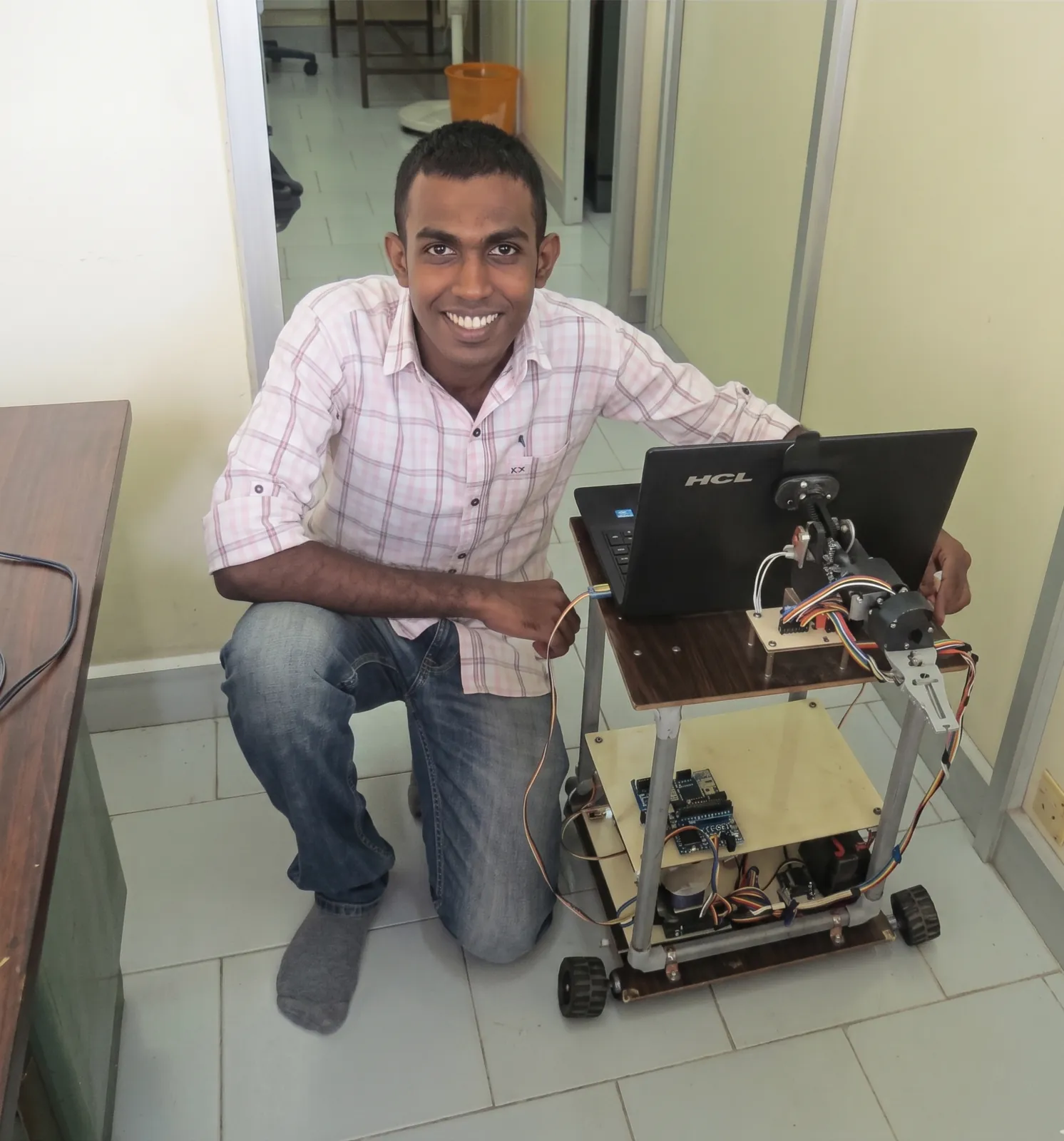 Leo Pauly kneeling beside CAMbot 1.0, a custom-built mobile robot carrying a laptop and a robotic manipulator.